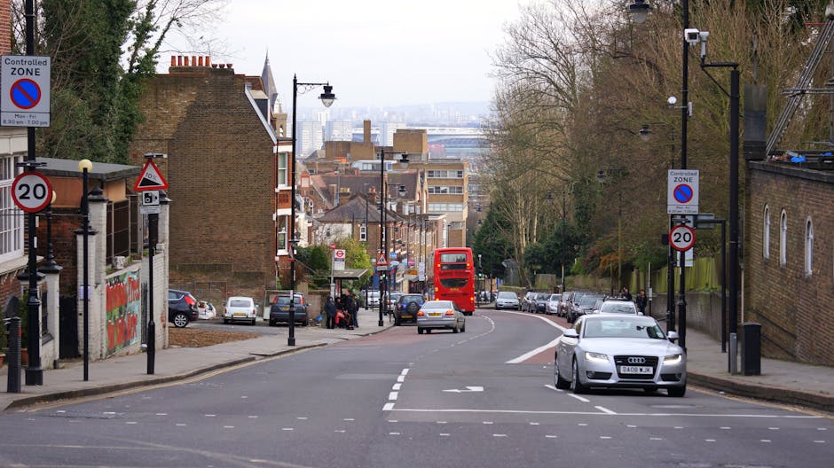 The image depicts a typical urban street scene with a gentle downward slope, featuring parked vehicles along both sides of the road including a silver Audi and several smaller cars, alongside a double